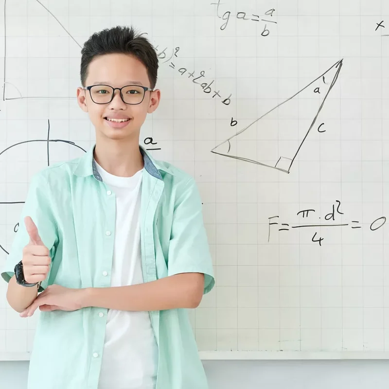sonriente-colegial-vietnamita-gafas-pie-pizarra-formulas-geometria-dibujos-mostrando-thumbs-up-min